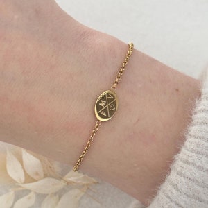 Könnte beinhalten: Ein goldenes Kettenarmband mit einem kleinen ovalen Anhänger, auf dem die Buchstaben "F", "M" und "V" in einem Kreuzmuster mit einem Herz in der Mitte angeordnet sind.