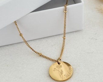 Op de afbeelding: Een gouden ketting met een ronde hanger met een delicaat bloemenmotief en de naam "Selina" erop gegraveerd.