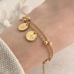 Könnte beinhalten: Ein goldfarbenes Armband mit zwei feinen Ketten und zwei runden Anhängern. Ein Anhänger zeigt ein Weizen-Design, der andere zwei Hände, die sich umfassen. Das Armband wird am Handgelenk getragen und zeigt sein elegantes Design.