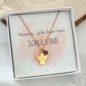 Könnte beinhalten: Eine goldene Engelkette mit einem kleinen Anhänger und dem Namen "Elena" darauf graviert. Die Kette befindet sich in einer weißen Box mit dem Text "Hurra, ich bin ein SCHULKIND" darauf.