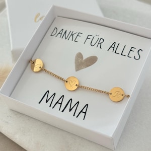 Può includere: Un braccialetto d'oro con tre ciondoli rotondi, uno con un cuore, uno con la lettera "L" e uno con la lettera "R". Il braccialetto è in una scatola bianca con un biglietto che dice "Danke für alles Mama".