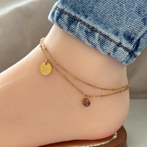 Könnte beinhalten: Ein goldenes Fußkettchen mit zwei Ketten und zwei Anhängern. Ein Anhänger ist eine runde Scheibe mit einem Sonnendesign. Der andere Anhänger ist eine runde Scheibe mit einem rosafarbenen Glitzerdesign.