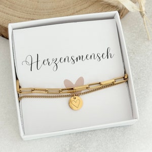 Könnte beinhalten: Ein goldenes Kettenarmband mit einem Herz-Anhänger in einer weißen Box mit dem Text "Herzensmensch" auf dem Deckel.