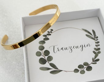 Op de afbeelding: Een gouden armband met de inscriptie "Trauzeugin" in een witte doos met een groen bloemmotief.