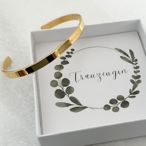 Könnte beinhalten: Ein goldenes Armreif-Armband mit der Inschrift "Trauzeugin" in einer weißen Box mit grünem Blumendesign.