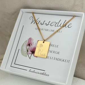 Peut inclure: Un collier pendentif rectangulaire doré avec un motif floral et le nom "Isabella" gravé. Le collier est présenté dans un coffret blanc avec du texte en allemand et une illustration florale.