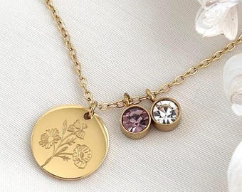 May include: A gold-colored necklace with a delicate chain. The necklace features a round pendant with a floral engraving and two small gemstone charms, one purple and one clear. The necklace is displayed on a white surface.