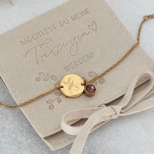 May include: A gold-colored bracelet with a small round pendant engraved with a floral design and the letter "S". A small purple gemstone is set next to the pendant. The bracelet rests on a beige pouch with text.
