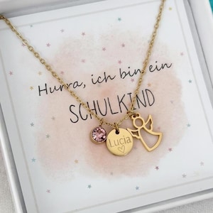 Könnte beinhalten: Eine goldfarbene Halskette mit feiner Kette und drei Anhängern: einem kleinen rosa Kristall, einer runden Scheibe mit der Gravur Lucia und einem Herz sowie einer Engelsfigur. Die Kette liegt auf einer Karte mit dem Text Hurra, ich bin ein SCHULKIND.