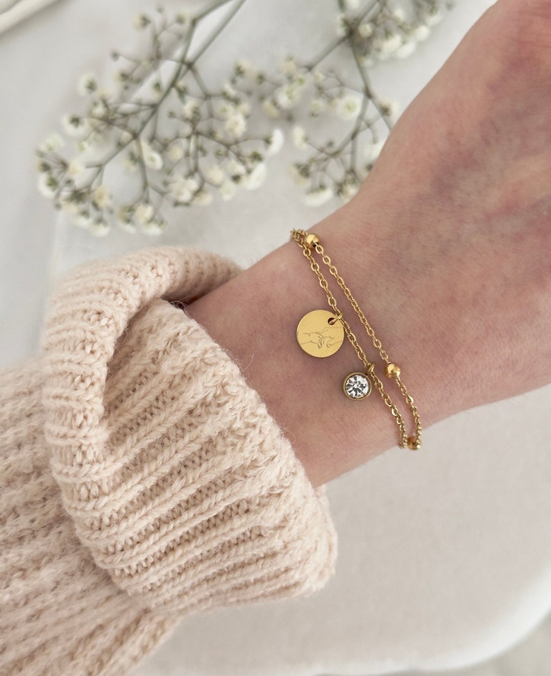 May include: A gold-colored double-strand bracelet with a round charm and a small, clear crystal. The charm has an engraved design. The bracelet is worn on a wrist, with a beige sweater in the background.