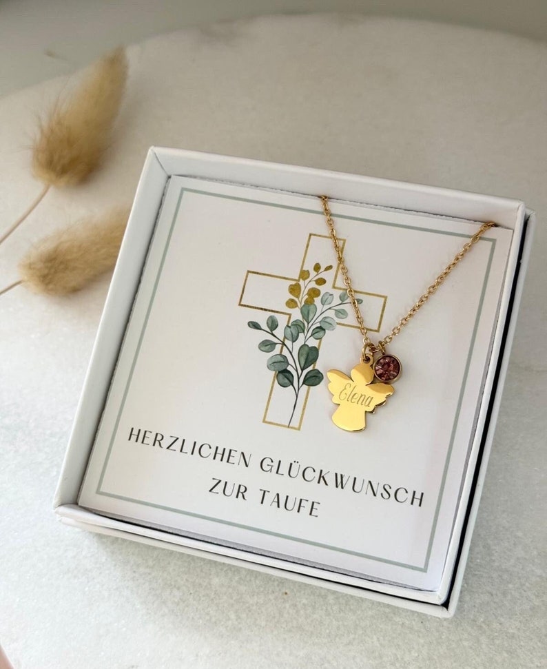 May include: A white gift box with a gold angel necklace inside. The necklace has a small pink gemstone and the name "Elena" engraved on the angel. The box has a white card with the text "Herzlichen Gl&uuml;ckwunsch zur Taufe" printed on it.