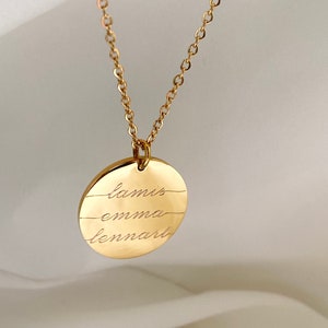 以下が含まれることがあります： 金色のネックレス。丸いペンダントには「lamis」「emma」「lennart」の3つの名前が刻まれています。チェーンも金色で、繊細なデザインです。名前は筆記体で刻まれ、ジュエリーにパーソナルなタッチを加えています。