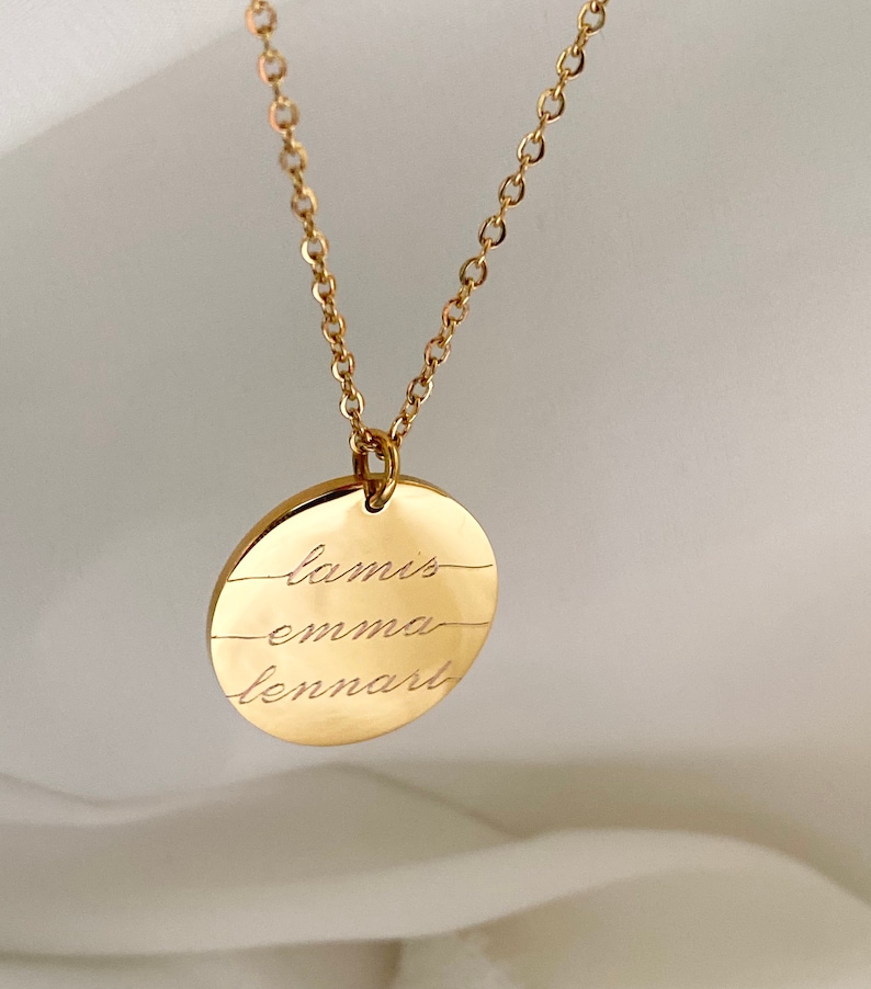 以下が含まれることがあります： 金色のネックレス。丸いペンダントには「lamis」「emma」「lennart」の3つの名前が刻まれています。チェーンも金色で、繊細なデザインです。名前は筆記体で刻まれ、ジュエリーにパーソナルなタッチを加えています。
