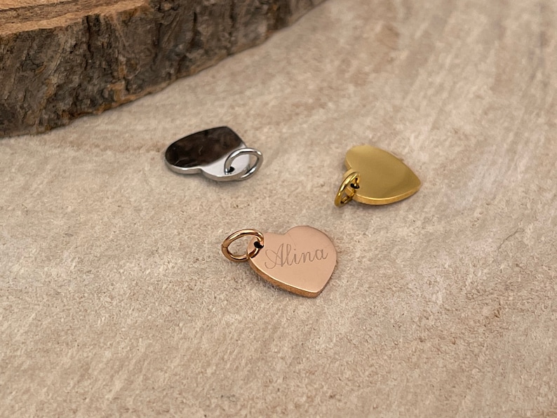 K&ouml;nnte beinhalten: Drei herzf&ouml;rmige Anh&auml;nger in Silber, Gold und Ros&eacute;gold. Der Ros&eacute;gold-Anh&auml;nger ist mit dem Namen "Alina" graviert. Jeder Anh&auml;nger hat eine kleine Schlaufe zum Befestigen an einer Kette oder einem Armband. Die Anh&auml;nger werden auf einer neutralen Oberfl&auml;che pr&auml;sentiert.