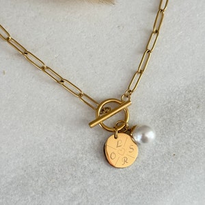 Könnte beinhalten: Goldkette mit einem Schiebeverschluss und einem runden Anhänger mit den Buchstaben L, O, S und R sowie einem Herzsymbol. Eine kleine weiße Perle hängt an dem Anhänger.