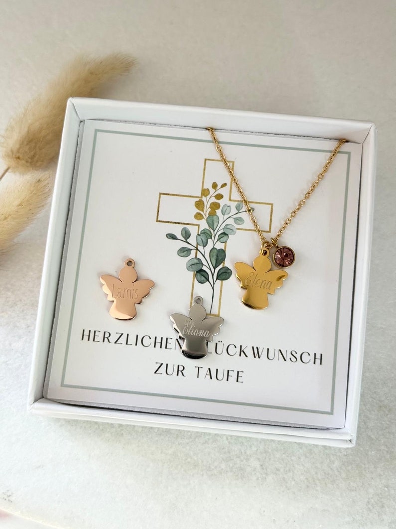 May include: A white box with a card inside. The card has a cross with green leaves and two angel pendants with the names "Elena" and "Liana" on them. The card also says "Herzlichen Gl&uuml;ckwunsch zur Taufe".