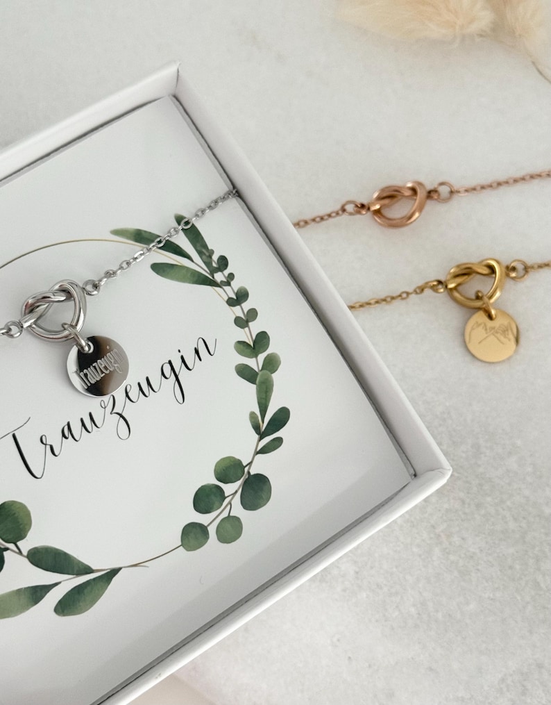 May include: Three delicate bracelets in silver, rose gold, and gold. Each features a heart-shaped knot design and a small, round charm. One bracelet is displayed inside a white box with the word "Trauzeugin" and a green leaf design.