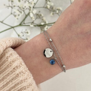 May include: A silver double-strand bracelet with a small round charm engraved with the name "Eliana" and a heart symbol. A blue gemstone pendant hangs below the charm. The bracelet is worn on a wrist.