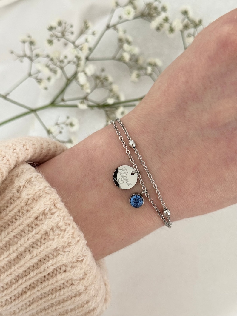 May include: A silver double-strand bracelet with a small round charm engraved with the name "Eliana" and a heart symbol. A blue gemstone pendant hangs below the charm. The bracelet is worn on a wrist.