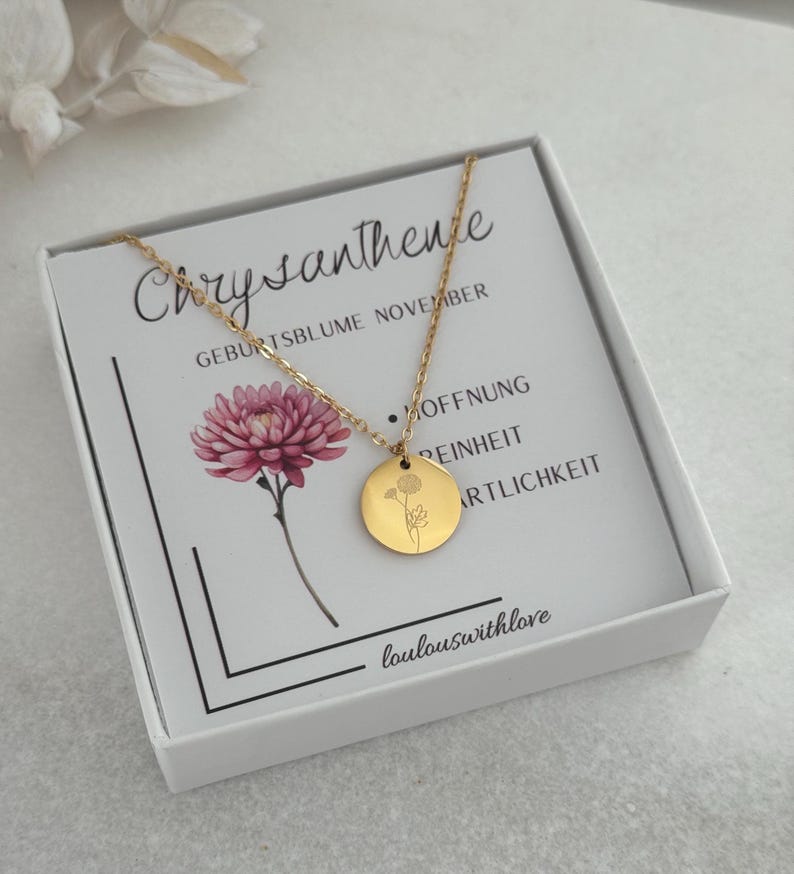 May include: A gold-colored necklace with a circular pendant featuring a floral design, presented on a card inside a white box. The card displays the word "Chrysantheme" and a pink flower illustration. The text "loulouswithlove" is visible.