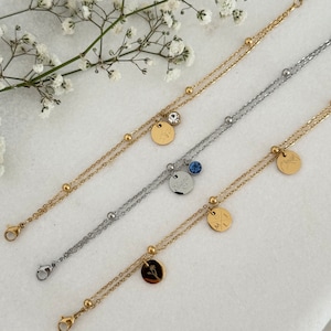 May include: Three layered bracelets in gold and silver tones. Each bracelet features a delicate chain design with small, round beads. The bracelets have small, round pendants with engraved designs and a small gemstone. The bracelets are displayed on a white surface.