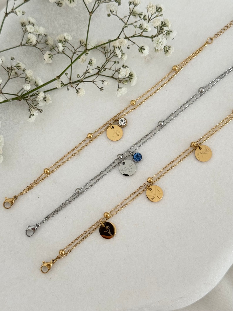 May include: Three layered bracelets in gold and silver tones. Each bracelet features a delicate chain design with small, round beads. The bracelets have small, round pendants with engraved designs and a small gemstone. The bracelets are displayed on a white surface.