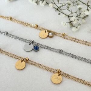 May include: Close-up of three layered necklaces. Two are gold-toned, one is silver-toned. Each necklace has a small, round pendant. One gold necklace has a clear crystal and a pendant with a bird design. Another has a pendant with initials.