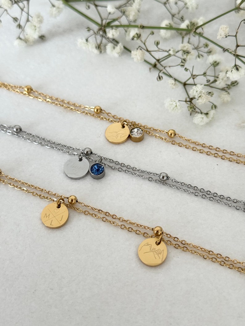 May include: Close-up of three layered necklaces. Two are gold-toned, one is silver-toned. Each necklace has a small, round pendant. One gold necklace has a clear crystal and a pendant with a bird design. Another has a pendant with initials.