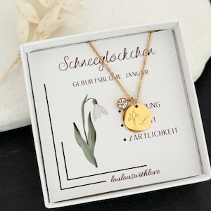 May include: A gold necklace with a small snowdrop charm and a larger charm with a snowdrop and the letter L. The necklace is in a white box with the text "Schneeglockchen Geburtsblume Januar" and "Z&auml;rtlichkeit" on it.