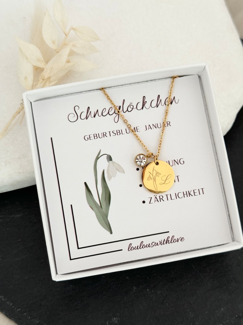 May include: A gold necklace with a small snowdrop charm and a larger charm with a snowdrop and the letter L. The necklace is in a white box with the text "Schneeglockchen Geburtsblume Januar" and "Z&auml;rtlichkeit" on it.