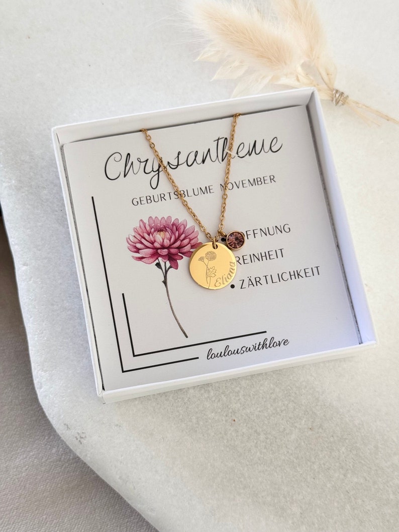 May include: A gold necklace with a round pendant that says "Elian" and a small pink crystal charm. The necklace is in a white box with a card that says "Chrysantheme Geburtsblume November" and lists the flower's meanings: "Offnung, Reinheit, Zartlichkeit".