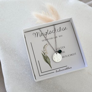 May include: A silver necklace with a round pendant featuring the letters "D", "R", "P", and "J" in a criss-cross pattern. The pendant has a small green gemstone in the center. The necklace is in a white box with a card that says "Maigl&ouml;ckchen Geburstsblume Mai" and "Gl&uuml;ck Liebe Artlichkeit" with the text "loulouswithlove" at the bottom.