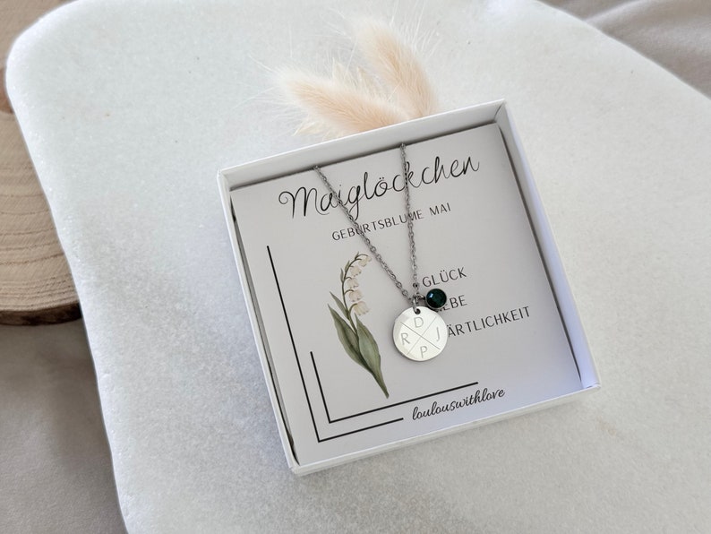 May include: A silver necklace with a round pendant featuring the letters "D", "R", "P", and "J" in a criss-cross pattern. The pendant has a small green gemstone in the center. The necklace is in a white box with a card that says "Maigl&ouml;ckchen Geburstsblume Mai" and "Gl&uuml;ck Liebe Artlichkeit" with the text "loulouswithlove" at the bottom.