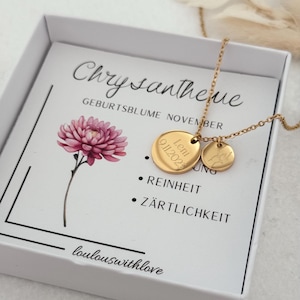 May include: A white box with a gold necklace inside. The necklace has two gold discs, one with the name "Leni" and the date "01.11.2021" engraved on it, and the other with a small flower design. The box also has a card with the text "Chrysantheme" and "Geburtsblume November" printed on it. The card also lists the meanings of the flower, "Reinheit" and "Z&auml;rtlichkeit". The bottom of the card reads "loulouswithlove".
