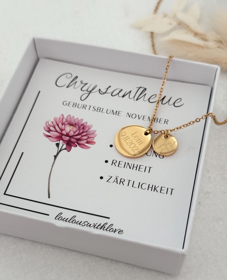 May include: A white box with a gold necklace inside. The necklace has two gold discs, one with the name "Leni" and the date "01.11.2021" engraved on it, and the other with a small flower design. The box also has a card with the text "Chrysantheme" and "Geburtsblume November" printed on it. The card also lists the meanings of the flower, "Reinheit" and "Z&auml;rtlichkeit". The bottom of the card reads "loulouswithlove".