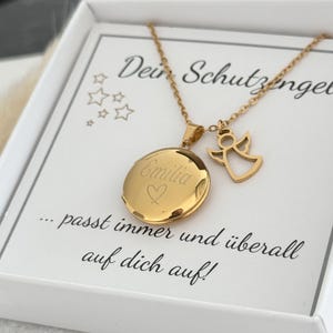 Könnte beinhalten: Goldene Halskette mit Anhänger in Form eines Medaillons und einem Engel. Das Medaillon ist mit dem Namen "Emilia" und einem Herz graviert. Die Halskette befindet sich in einer weißen Schachtel mit dem Text "Dein Schutzengel...passt immer und überall auf dich auf!"