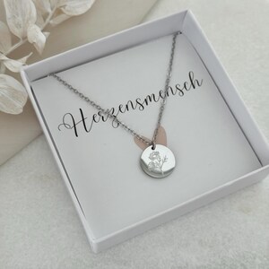 May include: A silver-colored necklace with a circular pendant featuring a floral design, presented in a white box. The necklace is delicate, with a fine chain. The box contains a card with the word "Herzensmensch" in cursive.