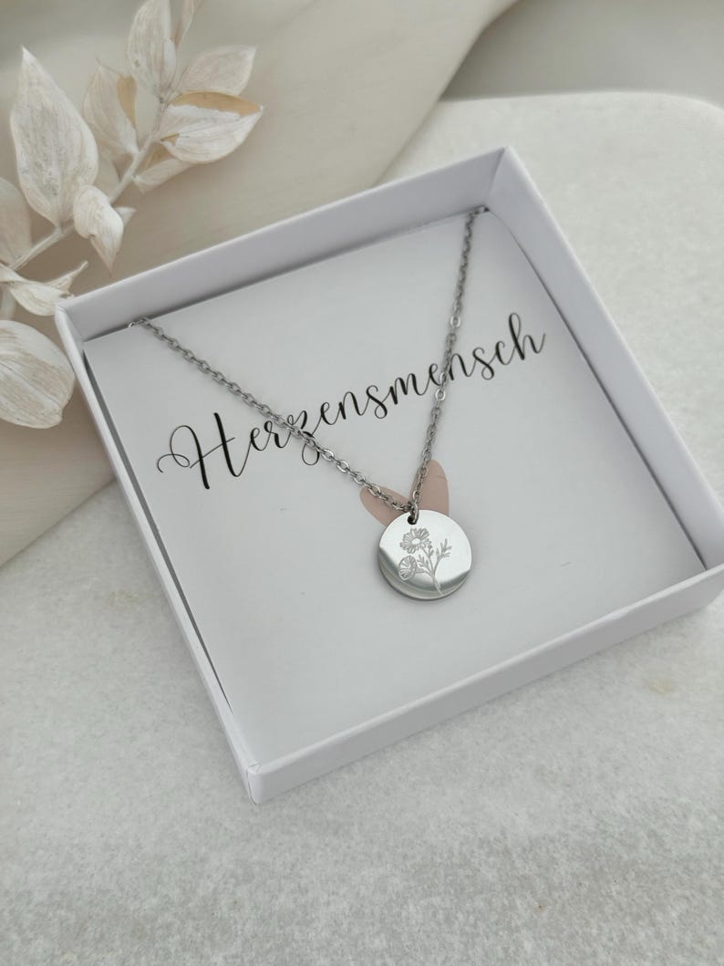 May include: A silver-colored necklace with a circular pendant featuring a floral design, presented in a white box. The necklace is delicate, with a fine chain. The box contains a card with the word "Herzensmensch" in cursive.