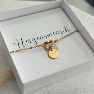 May include: A gold-colored bracelet with a knot design and a small circular charm. The charm features a mountain range engraving. The bracelet is presented in a white box with the word "Herzensmensch" printed on the inside.