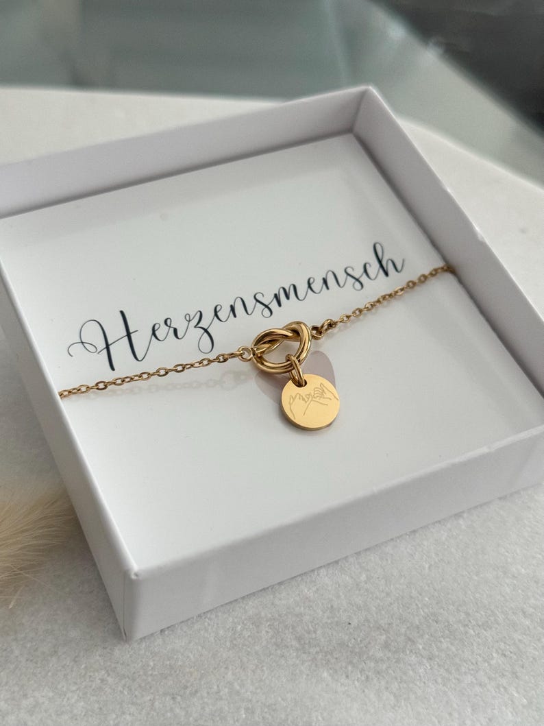 May include: A gold-colored bracelet with a knot design and a small circular charm. The charm features a mountain range engraving. The bracelet is presented in a white box with the word "Herzensmensch" printed on the inside.