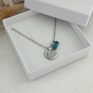 May include: A silver necklace with a round pendant featuring a floral design, accompanied by two blue gemstone charms. The necklace is presented in an open white box with the word "Couleurs" in gold script on the lid.