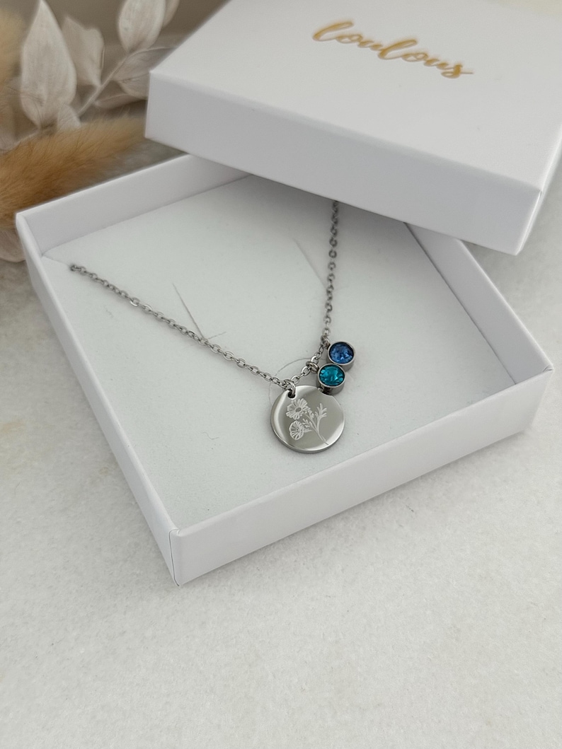 May include: A silver necklace with a round pendant featuring a floral design, accompanied by two blue gemstone charms. The necklace is presented in an open white box with the word "Couleurs" in gold script on the lid.
