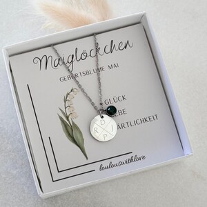 May include: A silver necklace with a round pendant engraved with initials and a small green gemstone. The necklace is presented in a white box with a card featuring floral illustrations and German text.