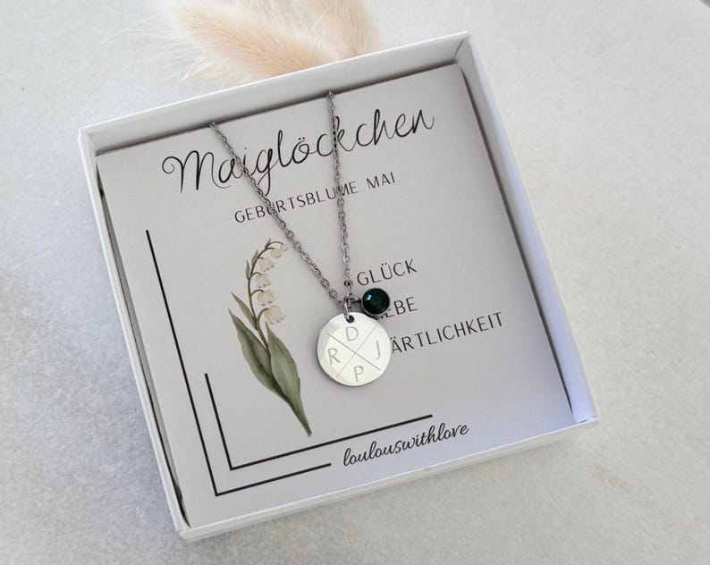 May include: A silver necklace with a round pendant engraved with initials and a small green gemstone. The necklace is presented in a white box with a card featuring floral illustrations and German text.
