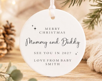 May include: A round white ceramic Christmas ornament hanging from a white ribbon. The ornament features black text that reads MERRY CHRISTMAS, Mummy and Daddy, SEE YOU IN 2027, and LOVE FROM BABY SMITH, accented with small four-pointed star icons.