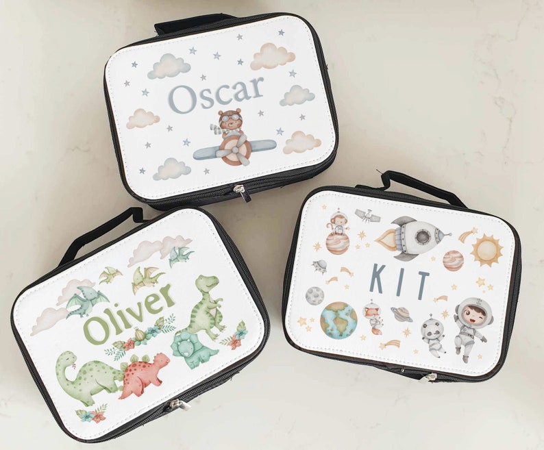 Puede incluir: Tres bolsas de almuerzo personalizadas con diferentes dise&ntilde;os. La primera bolsa tiene un dise&ntilde;o azul y blanco con nubes, estrellas y un oso piloto con el nombre "Oscar". La segunda bolsa tiene un dise&ntilde;o verde y blanco con dinosaurios y el nombre "Oliver". La tercera bolsa tiene un dise&ntilde;o blanco con planetas, estrellas, un cohete y un astronauta con el nombre "Kit".