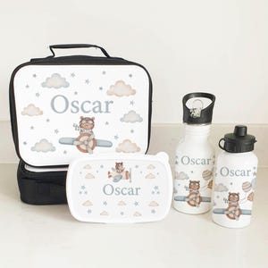 Puede incluir: Un juego de bolsa de almuerzo personalizado con un dise&ntilde;o blanco y azul que presenta un oso piloto volando en un avi&oacute;n. El juego incluye una bolsa de almuerzo, una fiambrera, una botella de agua y una botella de agua m&aacute;s peque&ntilde;a. El nombre "Oscar" est&aacute; impreso en cada art&iacute;culo.