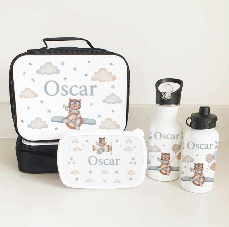 Puede incluir: Un juego de bolsa de almuerzo personalizado con un dise&ntilde;o blanco y azul que presenta un oso piloto volando en un avi&oacute;n. El juego incluye una bolsa de almuerzo, una fiambrera, una botella de agua y una botella de agua m&aacute;s peque&ntilde;a. El nombre "Oscar" est&aacute; impreso en cada art&iacute;culo.