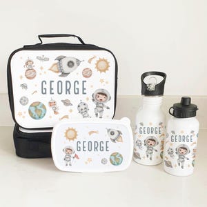 Puede incluir: Un juego de bolsa de almuerzo con tem&aacute;tica espacial con una bolsa de almuerzo aislada negra, una fiambrera blanca y dos botellas de agua blancas. La bolsa de almuerzo y la fiambrera presentan un cohete, planetas, estrellas y un astronauta. Las botellas de agua tienen el nombre "GEORGE" en ellas.