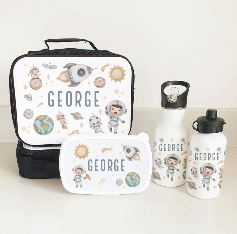 Puede incluir: Un juego de bolsa de almuerzo con tem&aacute;tica espacial con una bolsa de almuerzo aislada negra, una fiambrera blanca y dos botellas de agua blancas. La bolsa de almuerzo y la fiambrera presentan un cohete, planetas, estrellas y un astronauta. Las botellas de agua tienen el nombre "GEORGE" en ellas.